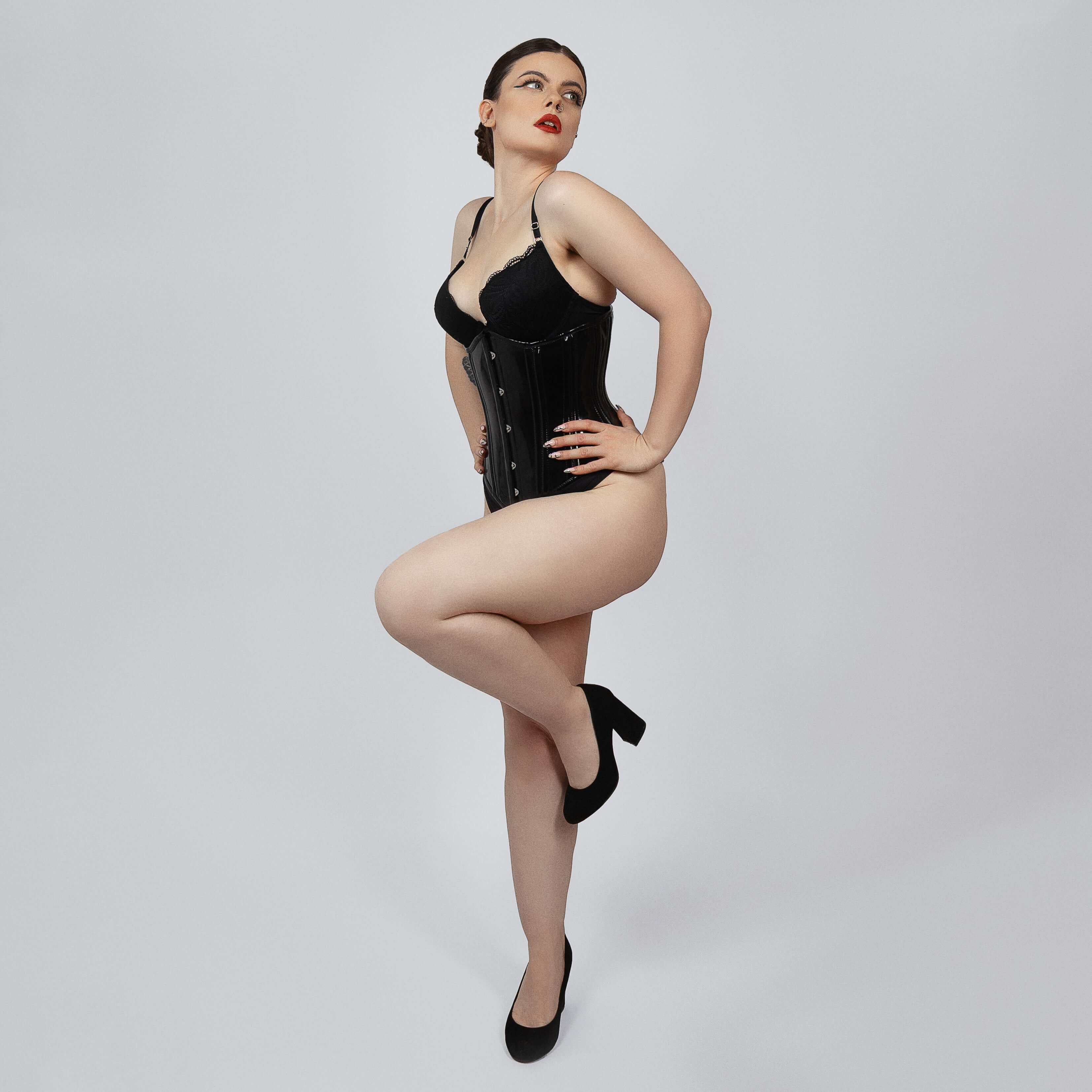 Model wearing black PVC underbust corset from side angle showing hourglass silhouette and hook and eye closure -- Daphne by Corsets Island
